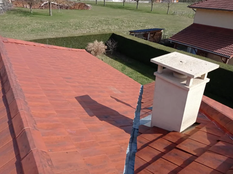 Vue en hauteur d’un toit en tuiles rouges avec une cheminée rectangulaire et un raccord métallique.
