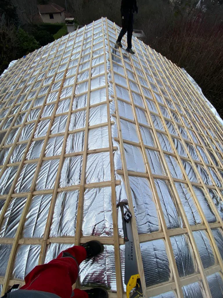 Vue d’une toiture en pente avec isolation réfléchissante, liteaux en bois et deux personnes au travail, dont une près d’un outil de mesure.